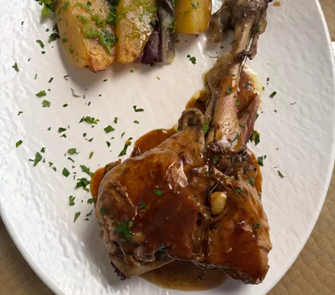 Una pieza de carne cubierta de salsa marrón oscura, con un hueso largo al descubierto y perejil picado por encima, sobre un plato blanco.