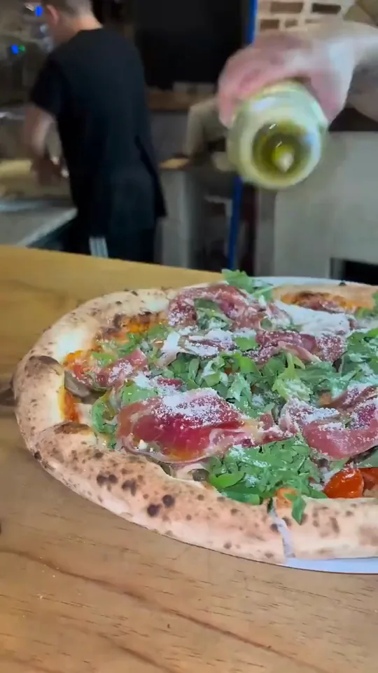 Una mano ralla queso por encima de una pizza de jamón serrano y rúcula, y después le da un hilo de aceite de oliva de lado a lado.
