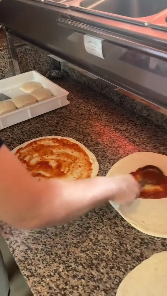 El cazo extiende la salsa de tomate en espiral sobre el disco de masa, empezando por el centro.
