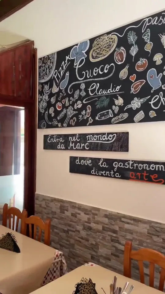 Nuestro comedor, vacío antes del servicio: en la pared, una gran pizarra pintada a tiza con dibujos de ingredientes y la palabra «Claudio», y debajo dos carteles negros escritos a mano, «Entra nel mondo da Marc» y «dove la gastronomia diventa arte». Delante, mesas con mantel de cuadros, sillas de madera y el zócalo de piedra.