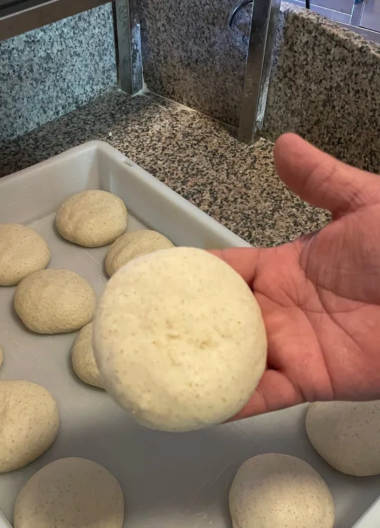 Una mano sostiene una bola de masa cruda sobre la bandeja blanca donde fermentan las demás, en nuestra encimera de granito moteado.