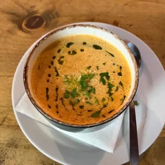 Un cuenco de cerámica de borde oscuro y rústico con una crema de color naranja intenso: por encima, gotas y trazos de aceite verde de hierbas, queso rallado y perejil picado. El cuenco está sobre un plato blanco, en una mesa de madera clara.